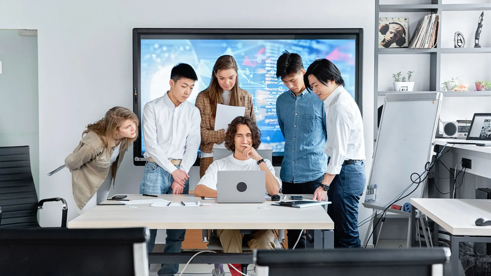 A group gathered around a laptop, discussing work in a bright office.